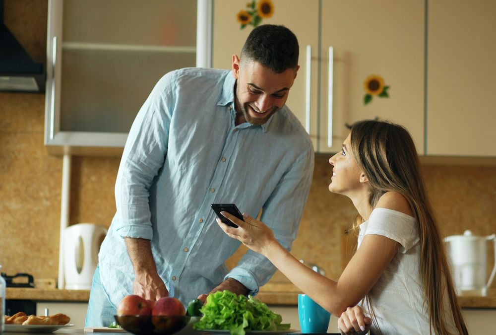 A couple smiling at a phone together in their kitchen
